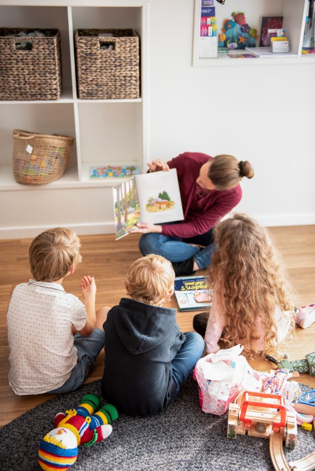Ella reading a book to her children while her children sit in front of her on the floor listening to the story. 