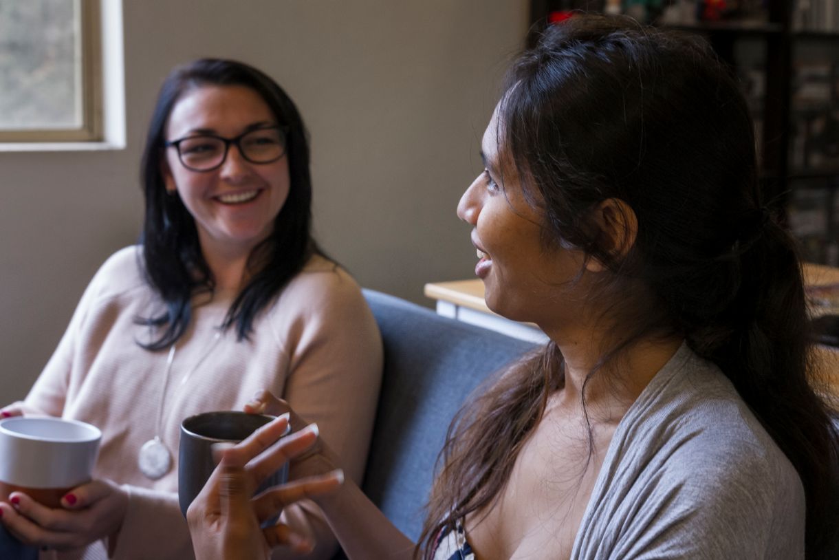Blair and her friend sitting on a couch holding mugs and talking.