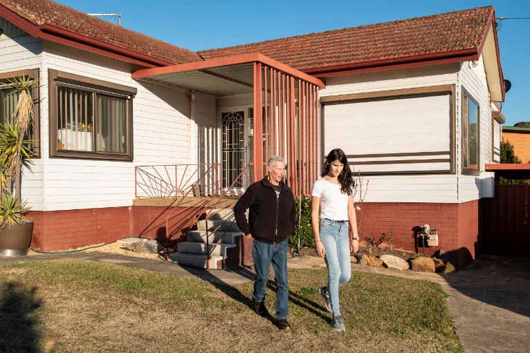 Garry pictured in front of his home while he walks away from the house with a woman next to him. They are both talking. 