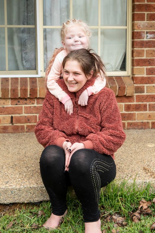 Kim and her daughter, Karma on their front porch. Karma behind Kim with her arms around Kim's shoulders. 