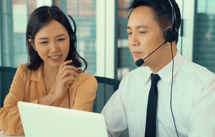 A man and a woman both wearing headsets in a business environment discussing work while looking at a computer.
